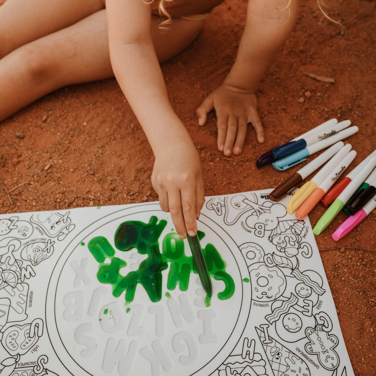 Child coloring a mandala on paper with markers on a red dirt surface