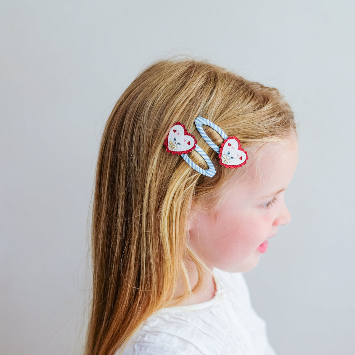 A young girl with long blonde hair is shown in profile wearing two heart-shaped hair clips. The clips have a light blue and white striped pattern with red borders and white embroidered faces.