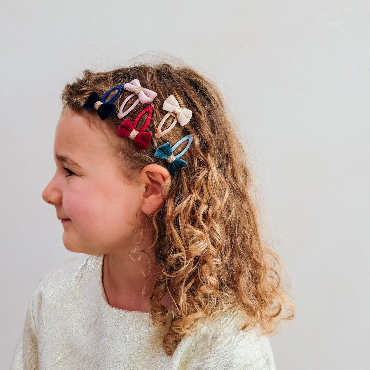A young girl with curly light brown hair is shown in profile wearing six colorful velvet bow hair clips in navy, pink, red, cream, and teal, set against a plain light gray background.