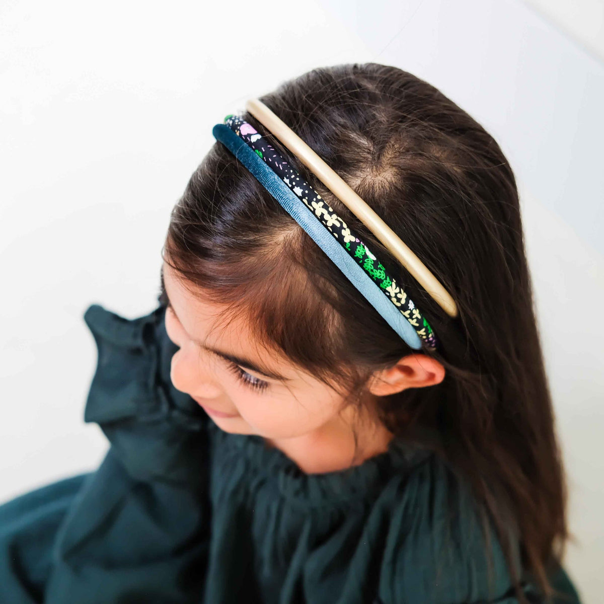A young girl with dark brown hair wearing a multi-layered headband. The headband features a light blue satin ribbon, a black and white leopard print ribbon, a solid beige ribbon, and a green glitter ribbon.