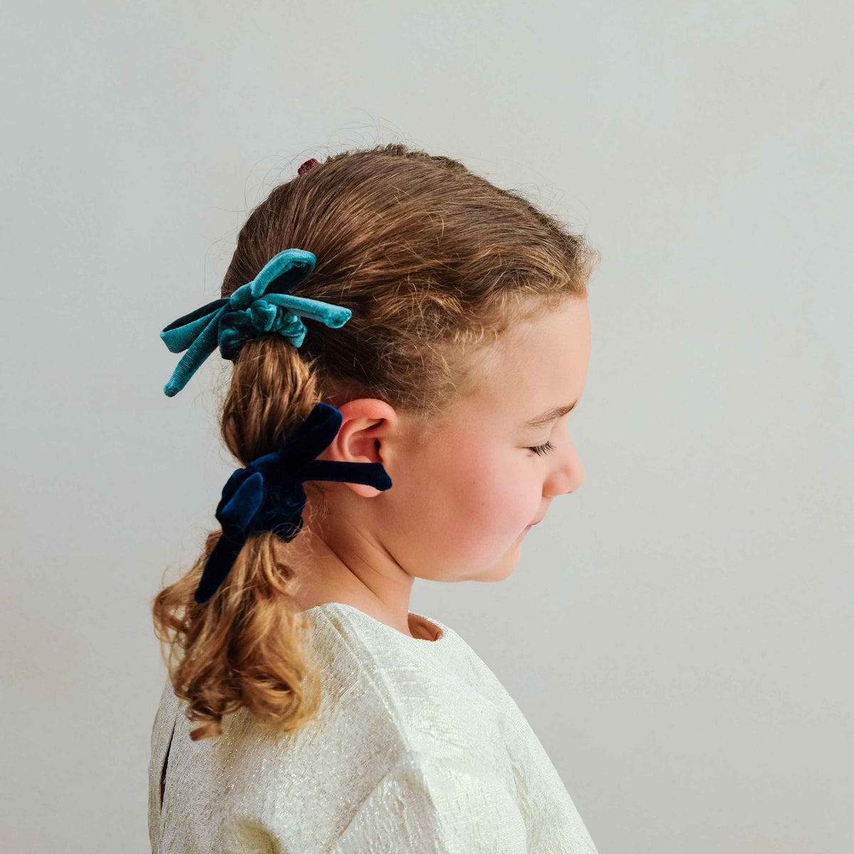 A young girl in profile wearing a white textured top, showcasing two velvet hair bows: a teal blue bow on top and a dark navy bow below, set against a plain light gray background.