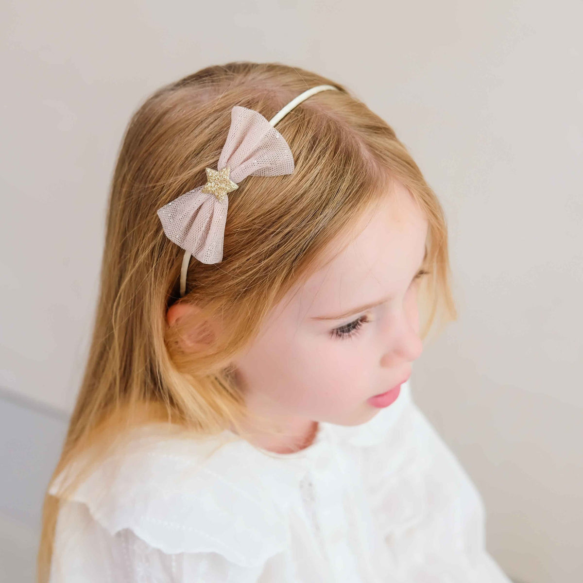 A young girl with long blonde hair wearing a light pink bow headband with a glittery gold star accent, paired with a white ruffled top against a soft beige background.
