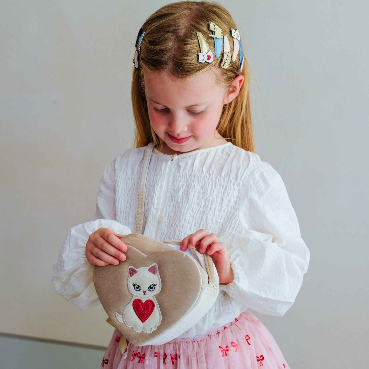 A young girl with blonde hair in a white smocked blouse and pink skirt holds a beige heart-shaped fabric bag with a white cat and red heart applique, set against a plain light gray background.