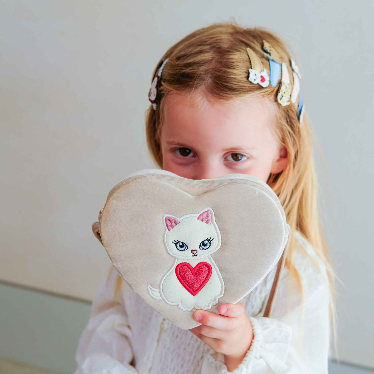 A young girl with blonde hair in a white dress holds a heart-shaped beige cushion or pillow in front of her face. The cushion features an embroidered white cat with a red heart on the front.
