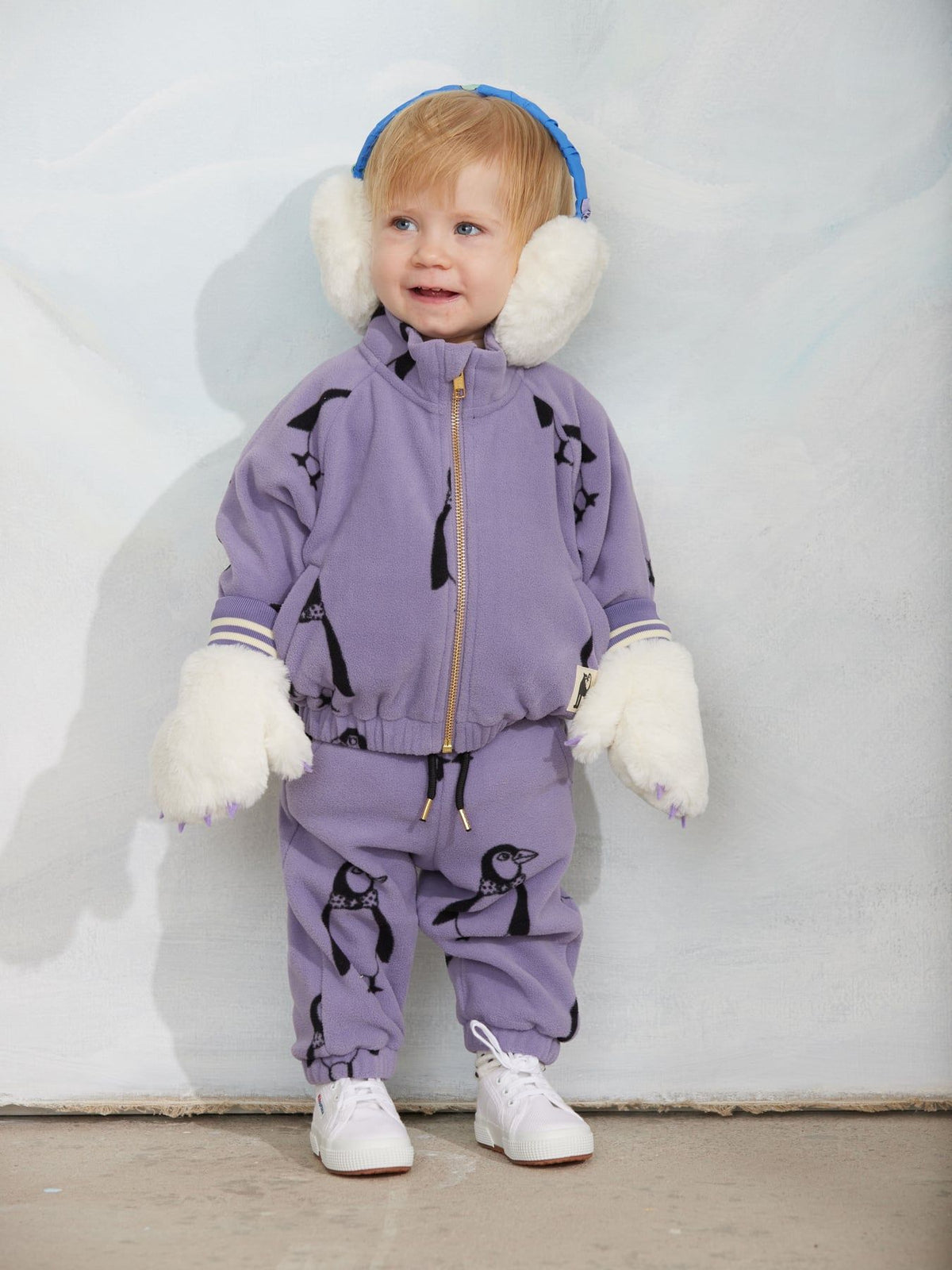 A toddler stands against a light gray wall wearing a lavender penguin-print zip-up jacket and matching pants, with white faux-fur mittens, white sneakers, and blue-and-white earmuffs.