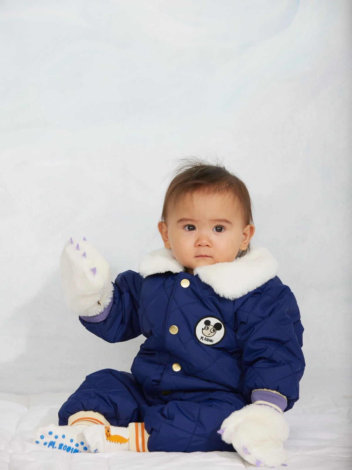 A baby wearing a white gloves and royal blue quilted baby snowsuit with a white faux-fur collar, gold snap buttons, striped cuffs and hem, and a cartoon patch on the chest.