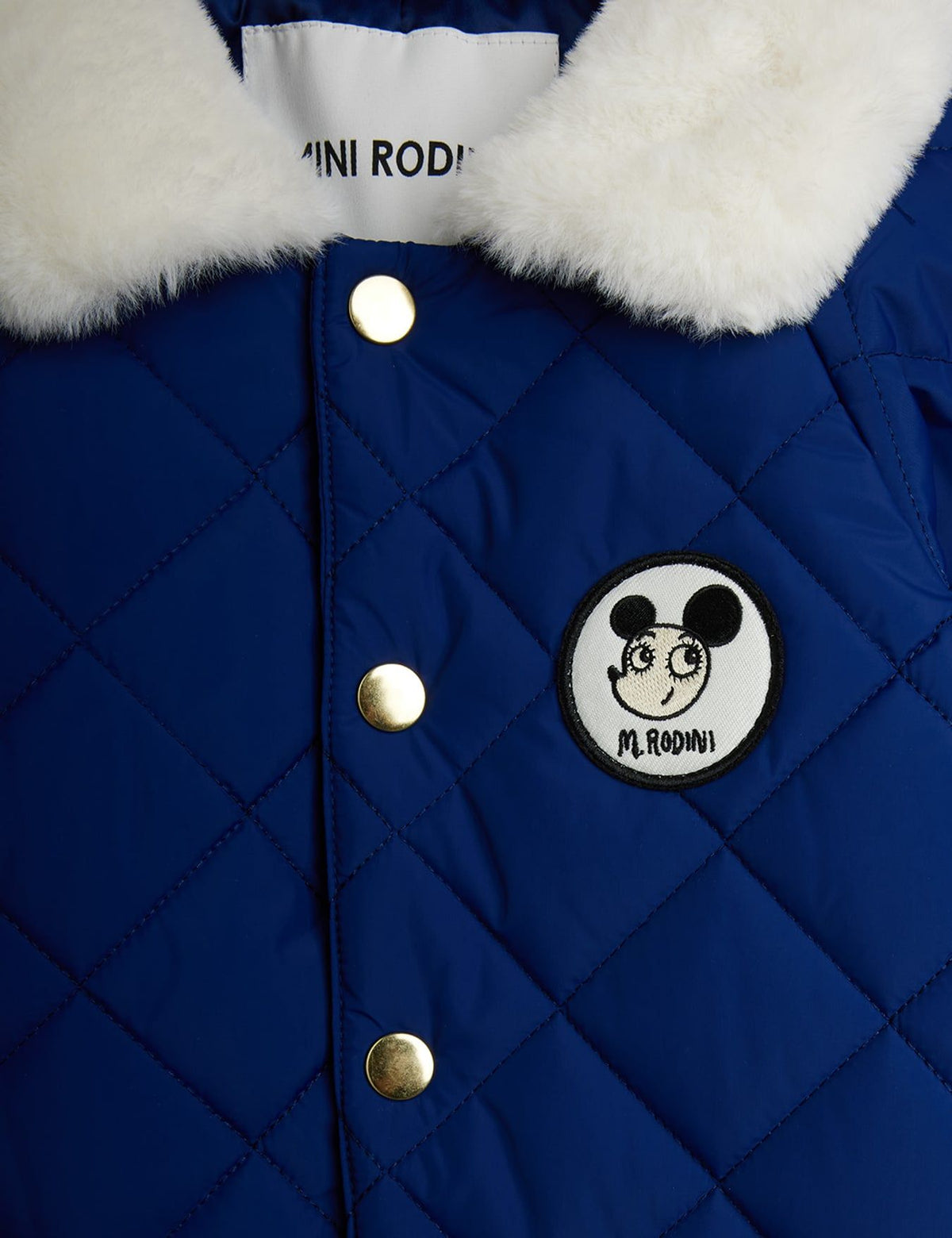 Close-up of a royal blue quilted jacket with a white faux-fur collar, gold-tone snap buttons, and a circular black-and-white patch featuring a cartoon mouse face and the text 'MINI RODI'.