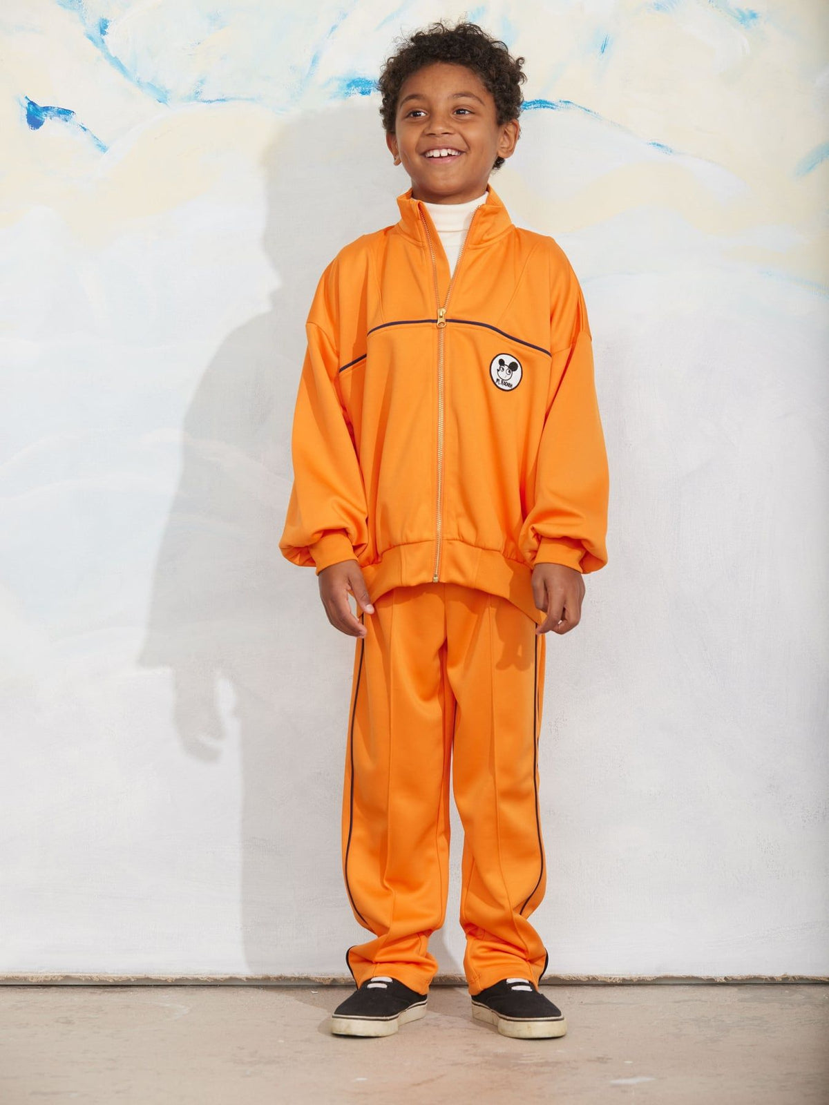 A young boy wearing a bright orange tracksuit with black piping details, standing against a white wall with a blue abstract painting in the background.