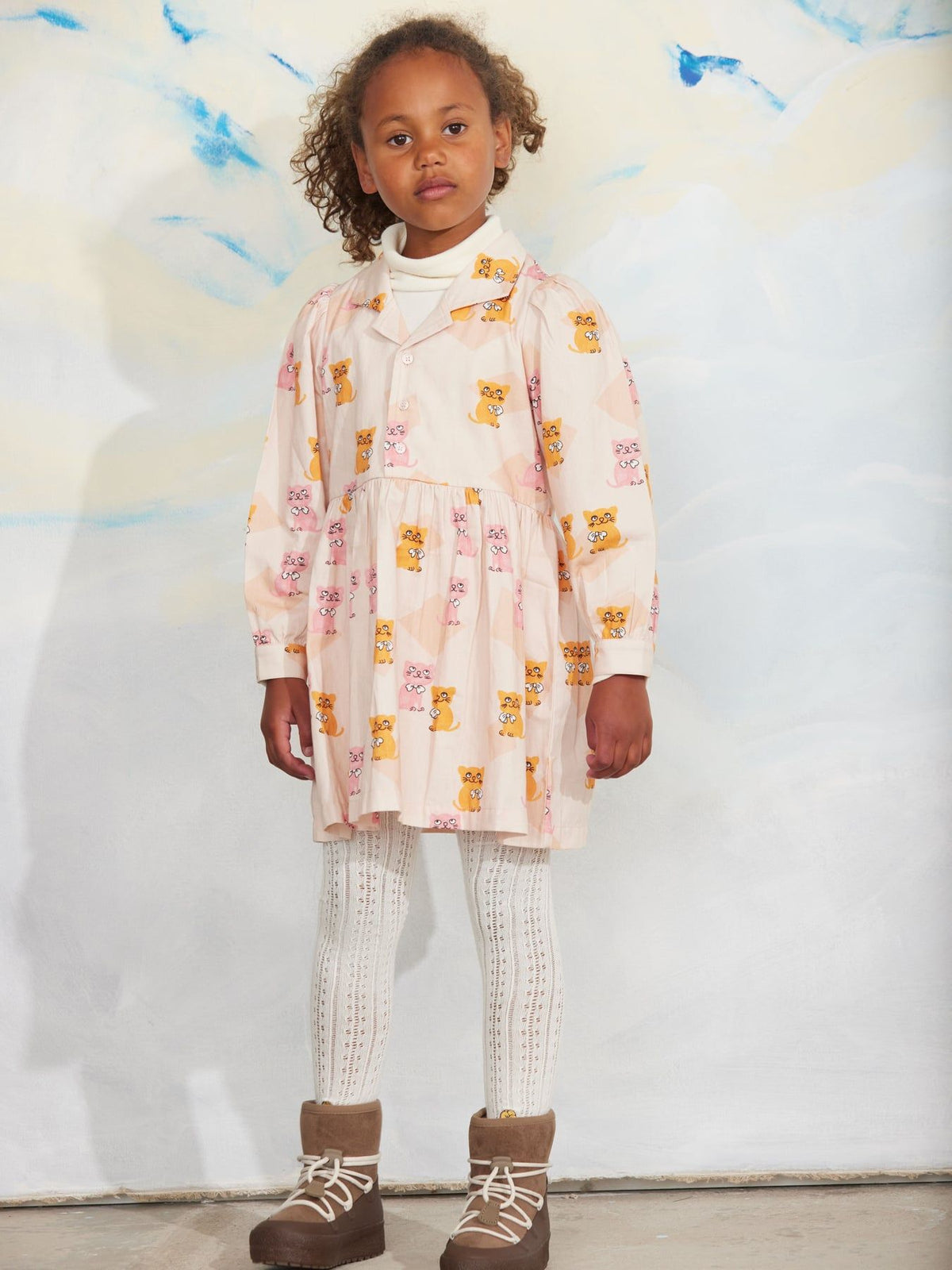 A young girl stands against a painted backdrop wearing a long-sleeve cream dress with a yellow and pink animal print, white knit tights, and brown lace-up boots.
