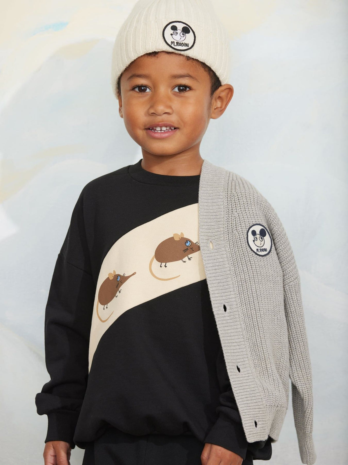 A young boy wearing a black and beige sweatshirt with mouse graphics, a light gray knit cardigan, and a cream beanie with a panda logo. The cardigan features a matching circular panda patch on the sleeve.