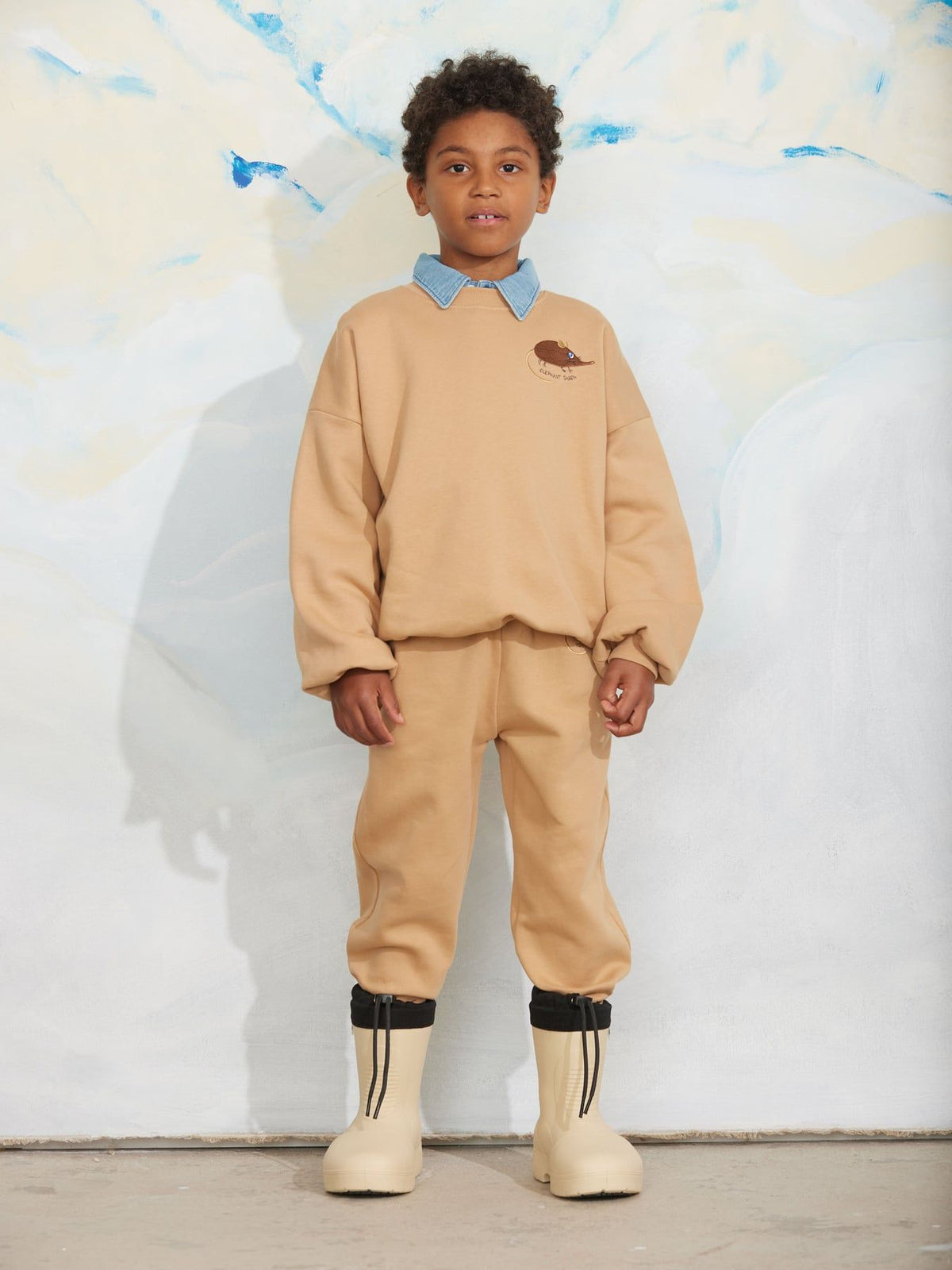 A child model wearing a beige sweatshirt and matching sweatpants with a light blue collar detail, black ankle cuffs, and beige boots, standing against a textured white wall.