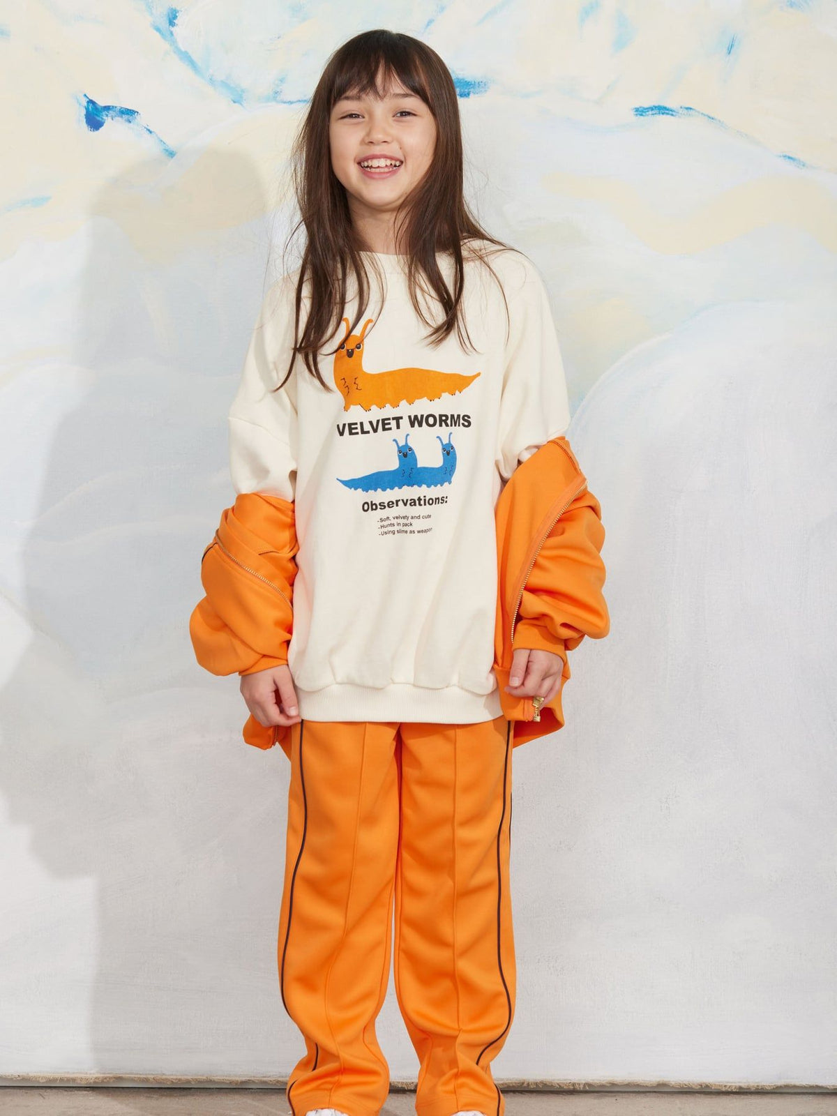 A young girl smiling while wearing a two-piece children's outfit from Velvet Worms, featuring an off-white sweatshirt with orange and blue whale graphics and matching orange puffer sleeves, paired with bright orange pants.