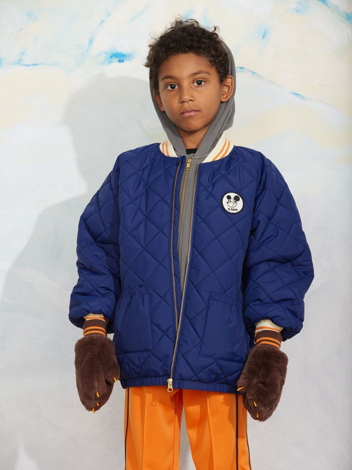 A child model wearing a navy blue quilted jacket with a gray hood, orange pants, and brown mittens, standing against a textured light gray and white background.