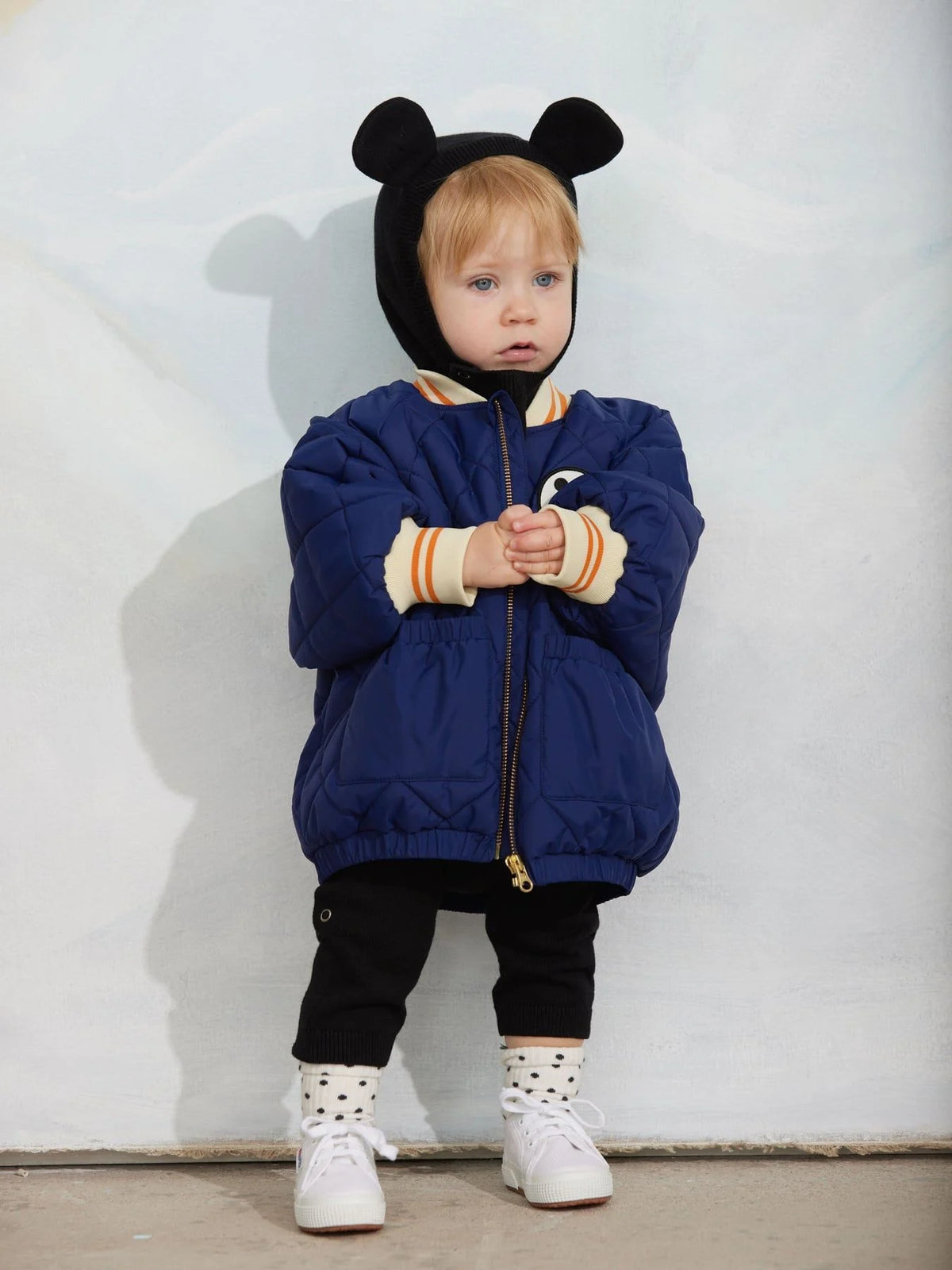 A toddler stands against a white wall wearing a navy blue quilted puffer jacket with a black hood featuring mouse ears, cream ribbed cuffs, black pants, and white sneakers.