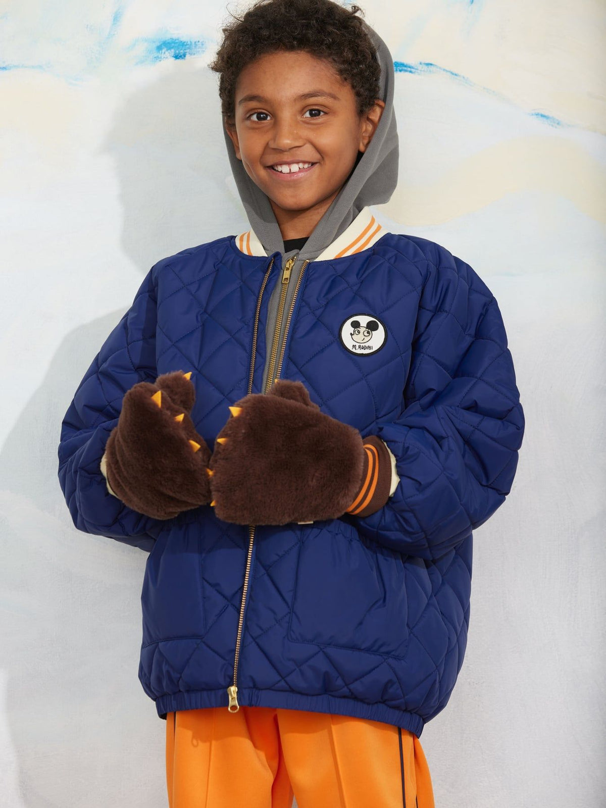 A smiling young boy wearing a blue quilted puffer jacket with a gray hood, brown fuzzy clawed gloves, and bright orange pants, standing against a light textured wall.
