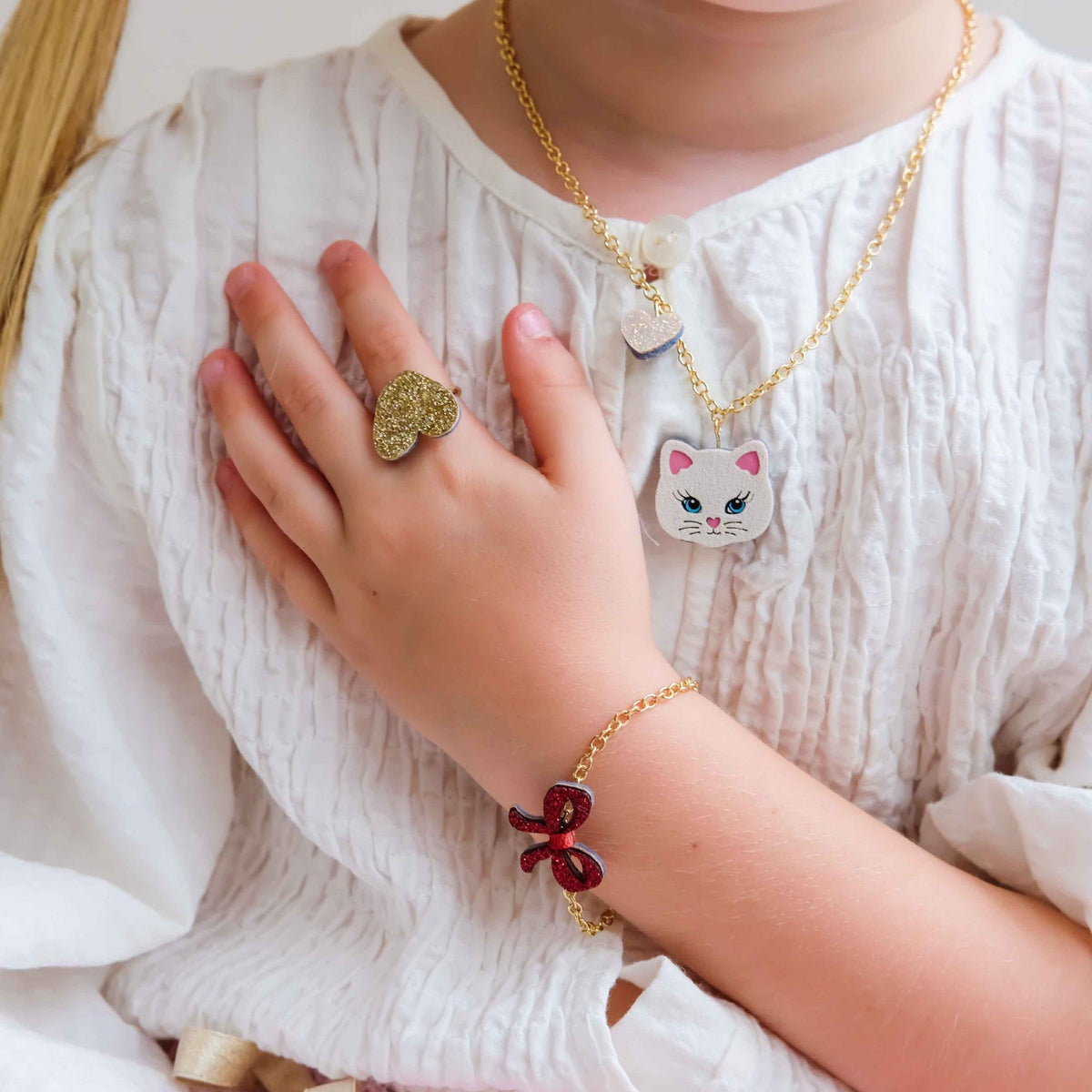 A close-up lifestyle shot of a child wearing a gold chain necklace with a white cat face pendant, a matching gold chain bracelet with a red bow charm, and a textured gold ring on their hand, all styled with a white crinkled blouse.