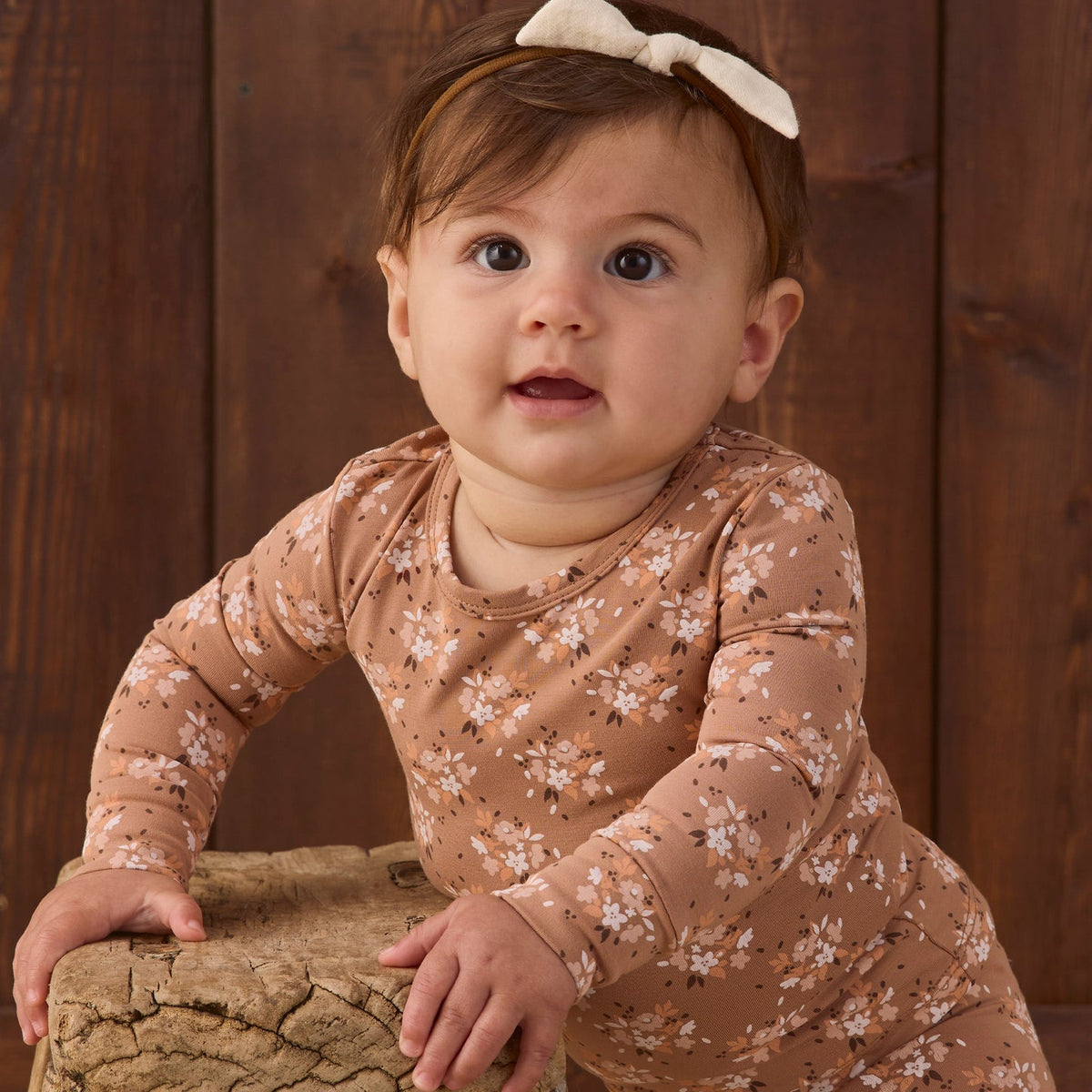 A baby girl wearing a long-sleeve tan jumpsuit with a white floral pattern and a matching cream bow headband, leaning on a rustic wooden stool against a dark wood plank background.