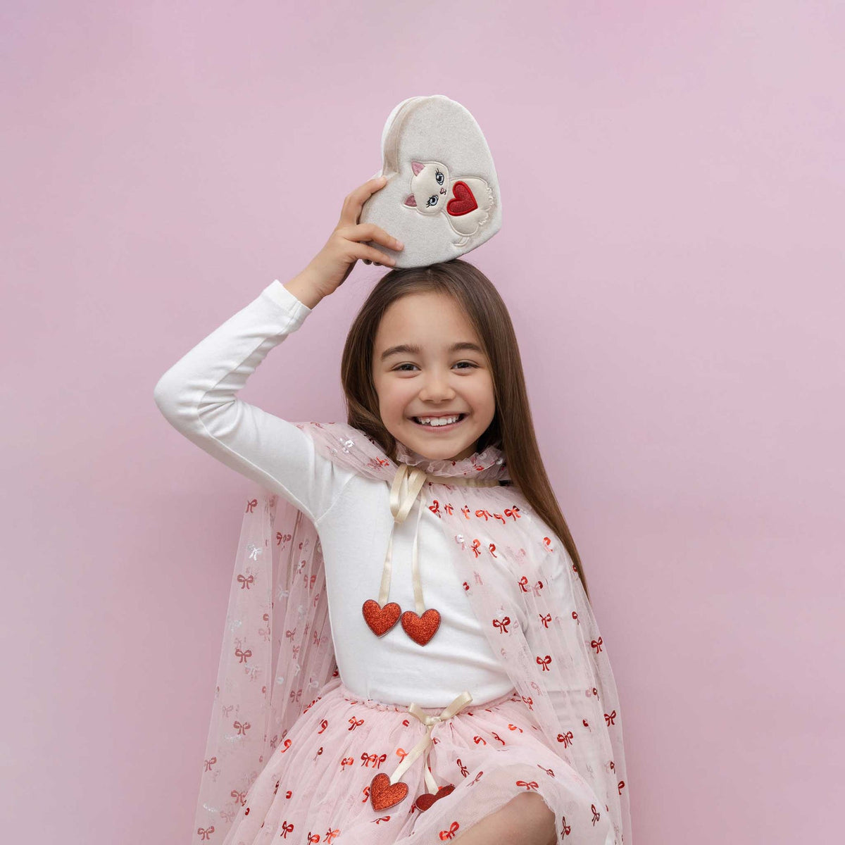 Young girl wearing a white shirt with red hearts and a pink dress, holding a white sandal with cartoon character on a pink background.
