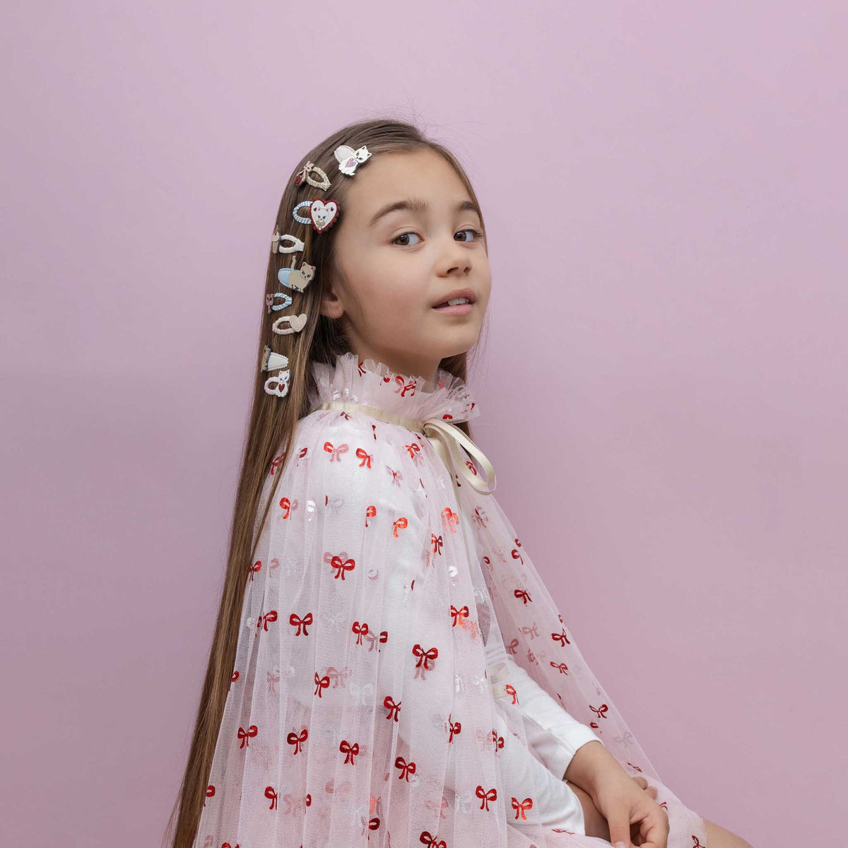 A young girl with long brown hair decorated with heart-shaped hair clips, wearing a white tulle dress with a red bow pattern, posed against a solid pink background.