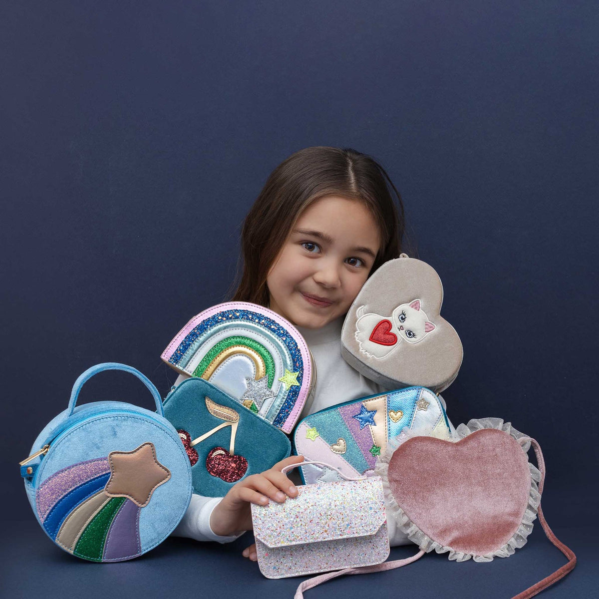 A young girl with long brown hair smiles while holding a collection of colorful, whimsical fabric bags and pouches against a dark navy blue background. The collection includes a light blue round bag with a rainbow and star, a teal bag with a mermaid tail and cherry, a light pink heart-shaped bag with a cat face, a glittery lavender clutch, and a dusty pink velvet heart pouch with a ruffled edge.