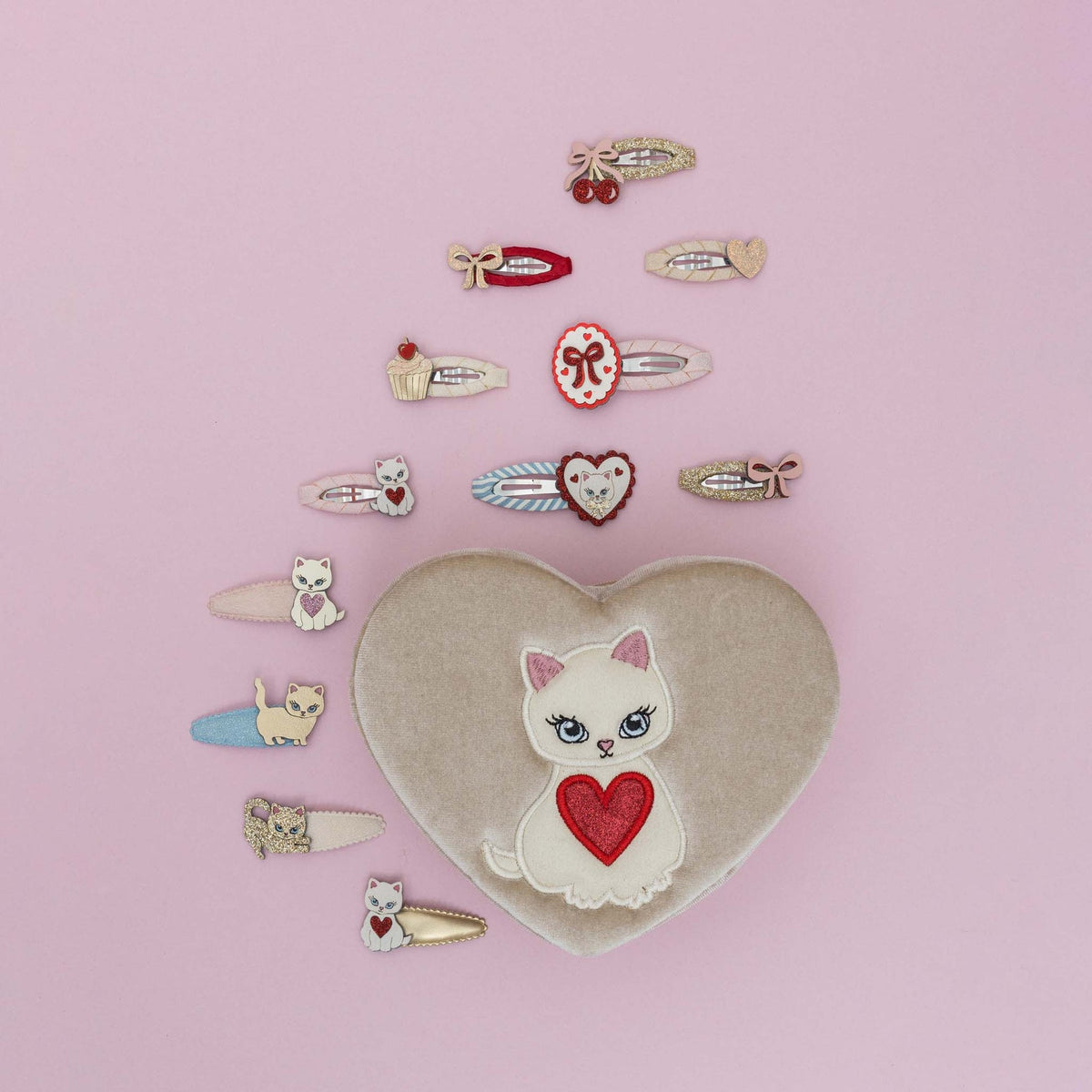 Heart-shaped pillow with cat design surrounded by various small hair accessories on a pink background