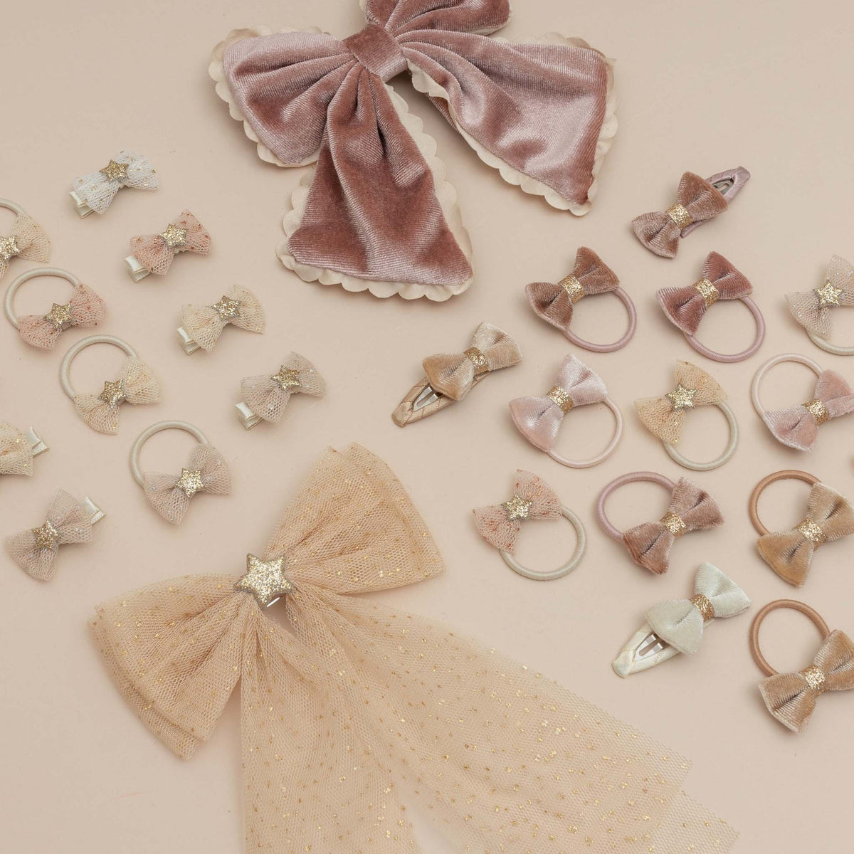 A flat-lay collection of pastel hair accessories on a solid beige background, including velvet and tulle bows, hair clips, and hair ties in soft pink, beige, and light blue tones, accented with glittery star details.