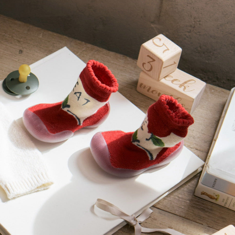 Red baby shoes with white soles on a wooden surface with blocks and a pacifier.