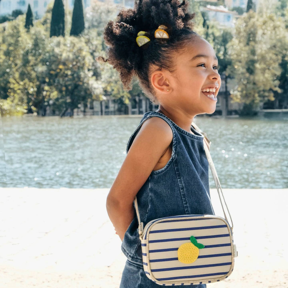 Child with a striped bag featuring a lemon design, standing by a lake.