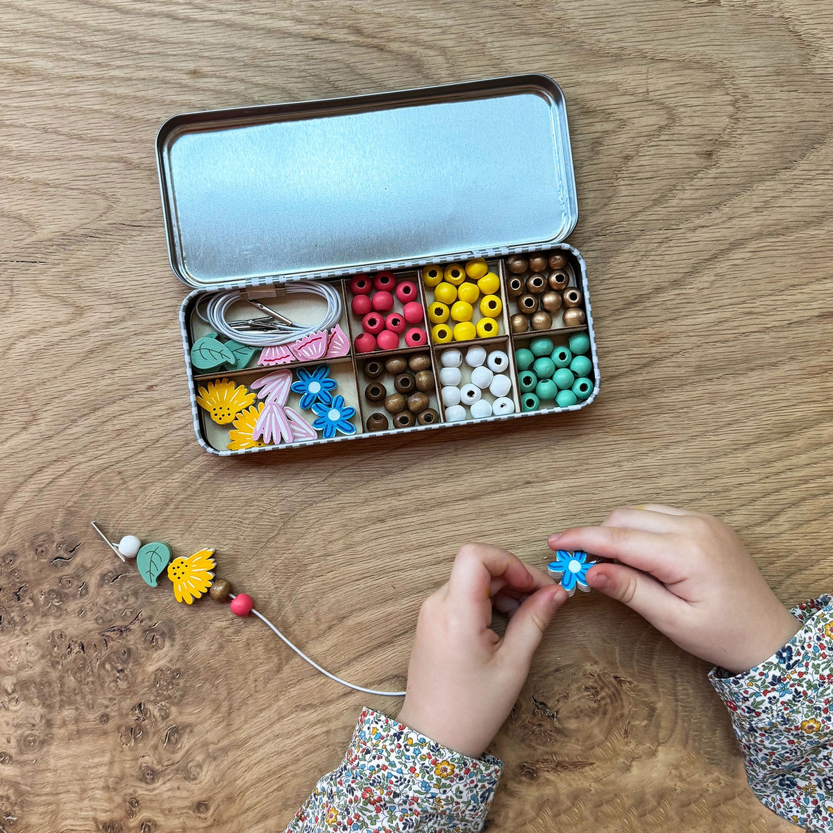Children's hands working with colorful beads on a wooden table, with a tin of beads open in the background.