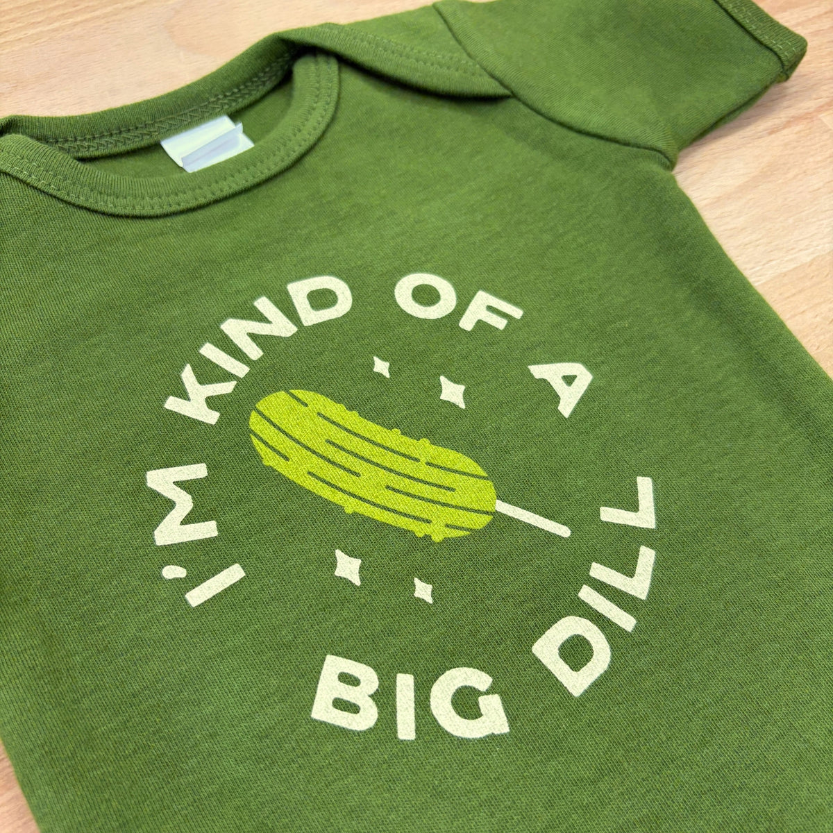 An olive green baby bodysuit laid flat on a light wood surface, featuring a white graphic print of a pickle and the text 'I'M KIND OF A BIG DILL'.