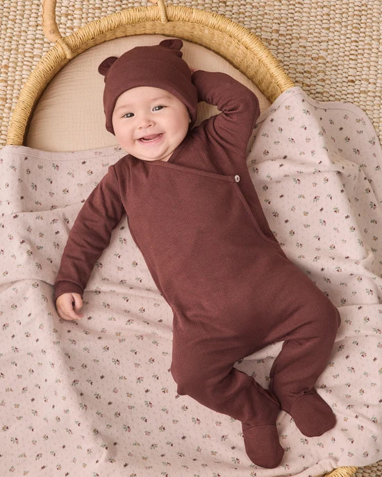 A smiling baby lies on their back on a patterned blanket inside a woven bassinet, wearing a brown footed sleeper and matching bear-ear hat.