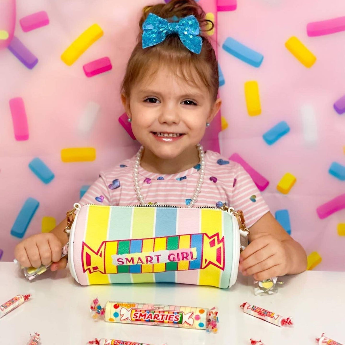Child holding a colorful pencil case with 'Smart Girl' text against a pink background with candy decorations.