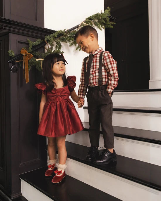 A young girl in a red ruffled dress and red-and-white plaid shirt stands beside a boy in brown suspenders and a matching plaid shirt on a black-and-white staircase, holding hands under a green garland.