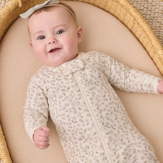 A baby lying on their back inside a woven Moses basket, wearing a cream-colored long-sleeve footless onesie with a delicate floral pattern and a matching bow headband.