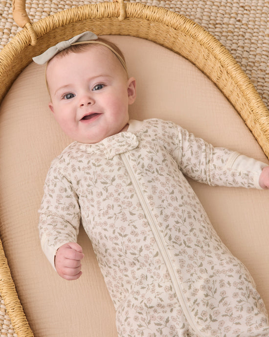 A baby lying on their back inside a woven Moses basket, wearing a cream-colored long-sleeve footless onesie with a delicate floral pattern and a matching bow headband.