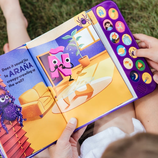 A child holding open "What Does Ñ Sound Like?: A Spanish-English Sound Book," showing an interior illustrated page featuring a purple animated character, a living room scene, and the text "la ARAÑA" on the left, with a vertical panel of sound buttons on the right edge.