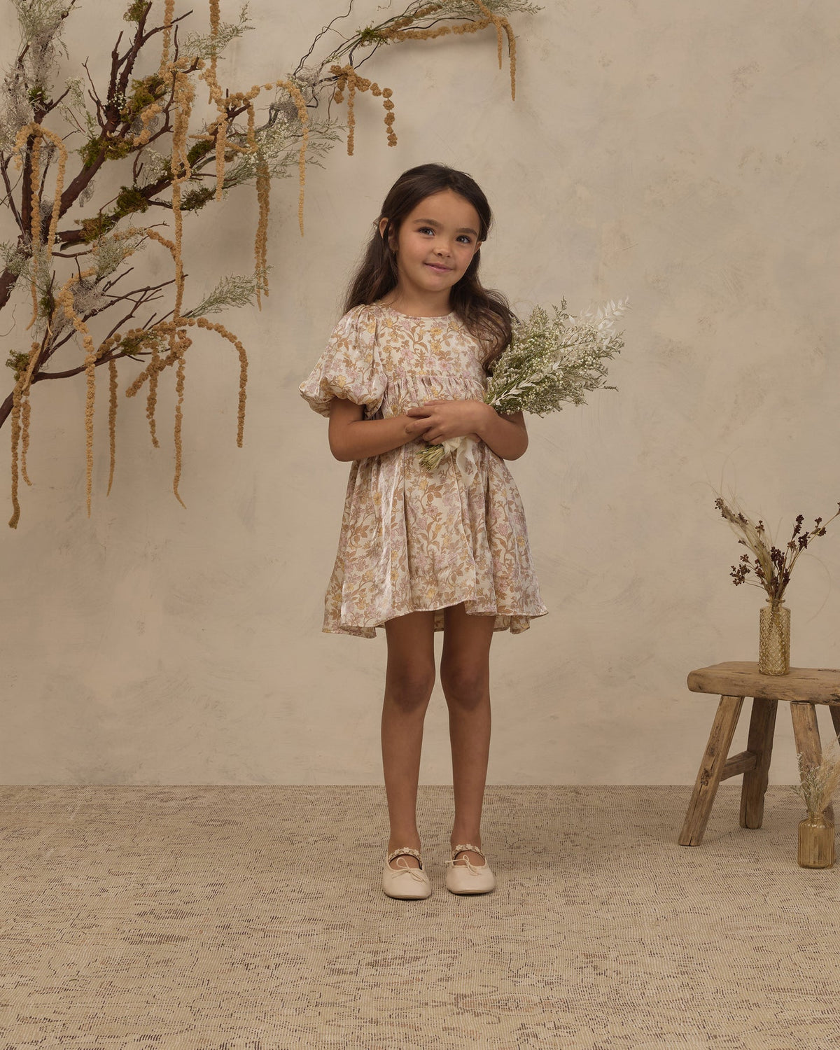 A young girl stands in a studio setting wearing a short, flutter-sleeve dress with a beige and yellow floral pattern. She is holding a small bouquet of dried white flowers and standing next to a rustic wooden stool with a vase of dried branches against a textured beige wall.