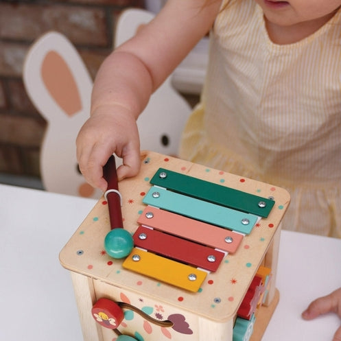 A toddler in a light pink dress plays with the wooden activity cube by Mentari, using a red mallet to tap the colorful xylophone on top.