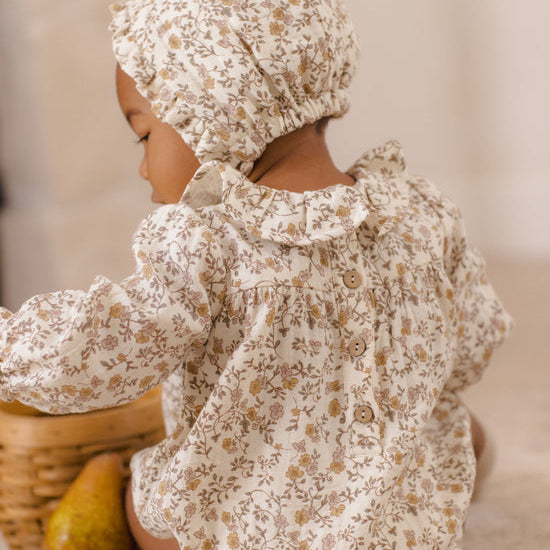 A baby wearing a cream-colored floral romper and matching bonnet, sitting on a carpeted floor next to a wicker basket and a pear.