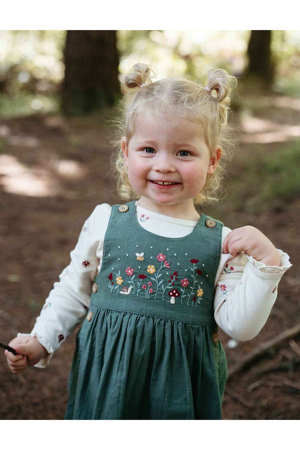 A young toddler girl with blonde pigtails smiles outdoors in a forest while wearing a dark green pinafore dress over a white long-sleeve shirt. The dress features colorful embroidered flowers, mushrooms, and small animals on the chest.