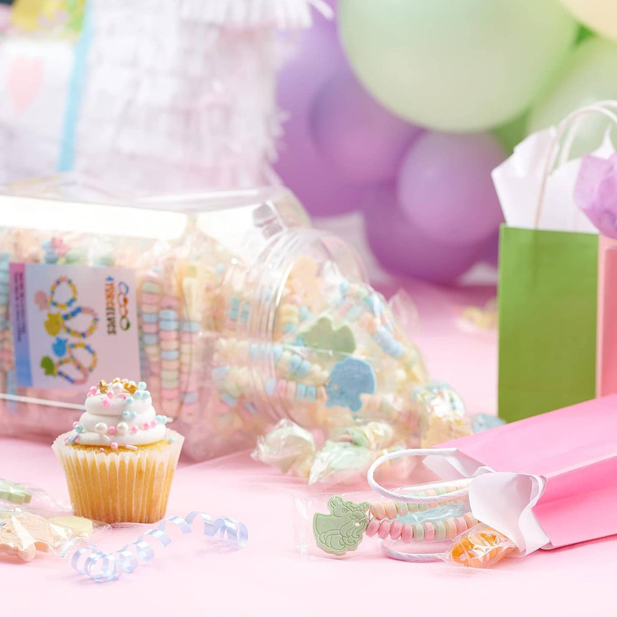 A styled tabletop scene with pastel party decorations, including balloons, a pink gift bag, a cupcake, and several clear jars and bags filled with colorful candy and wrapped sweets on a pink surface.