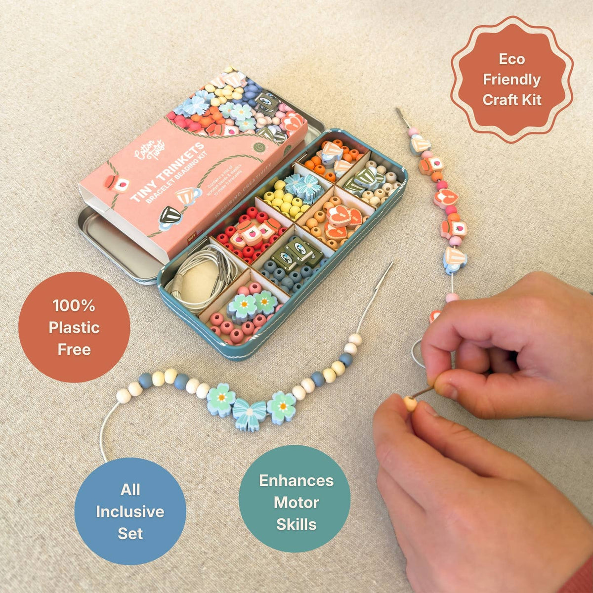 A bead craft kit shown on a light beige fabric background, with an open teal tin of colorful beads, a pink branded box, and a beaded cord being threaded by hands. Text callouts read 'Eco Friendly Craft Kit,' '100% Plastic Free,' 'All Inclusive Set,' and 'Enhances Motor Skills.