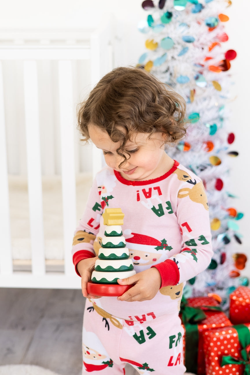 Child in festive pajamas holding a Christmas tree toy in front of a decorated Christmas tree.