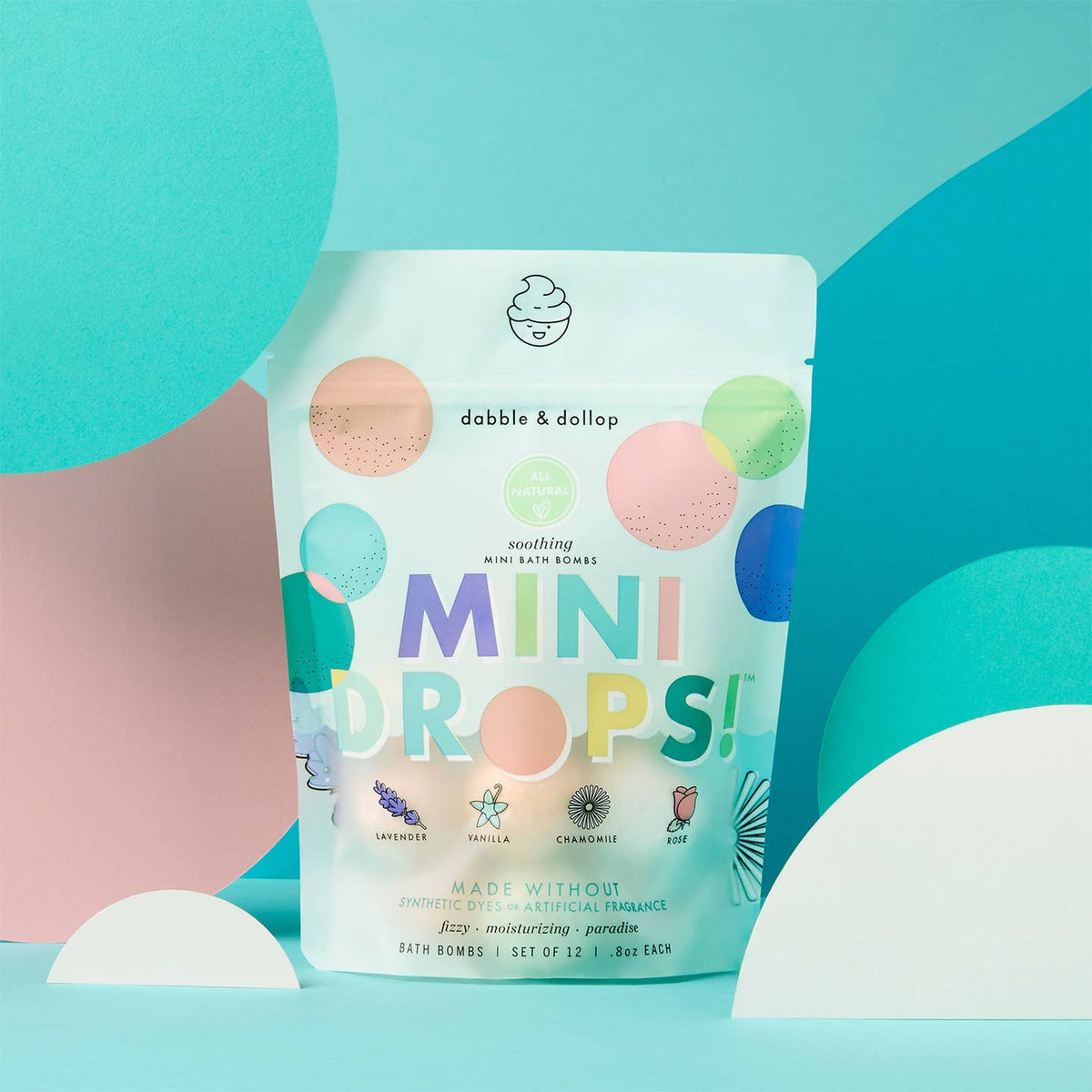Package of 'Mini Drops' bath bombs with colorful design on a blue and pink abstract background