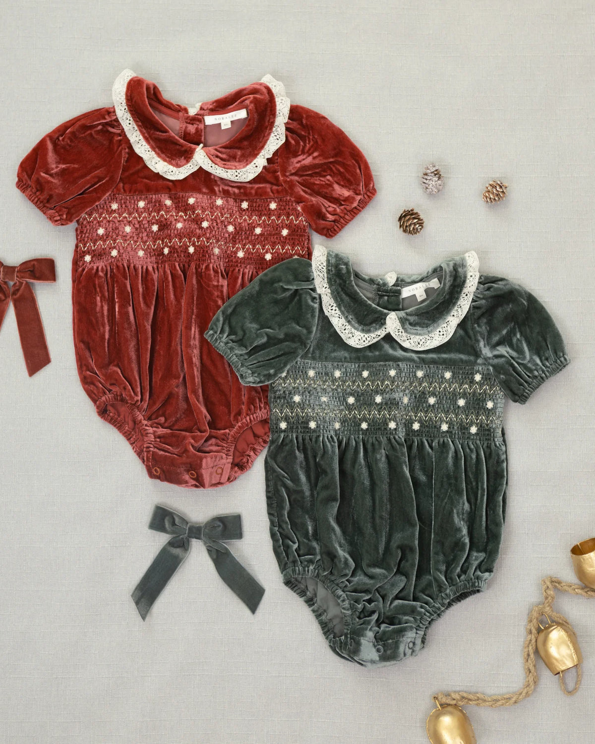 Two short-sleeve baby bloomers in velvet, one in deep red and one in dark green, laid flat on a light gray background with decorative bows, pinecones, and gold holiday lights.