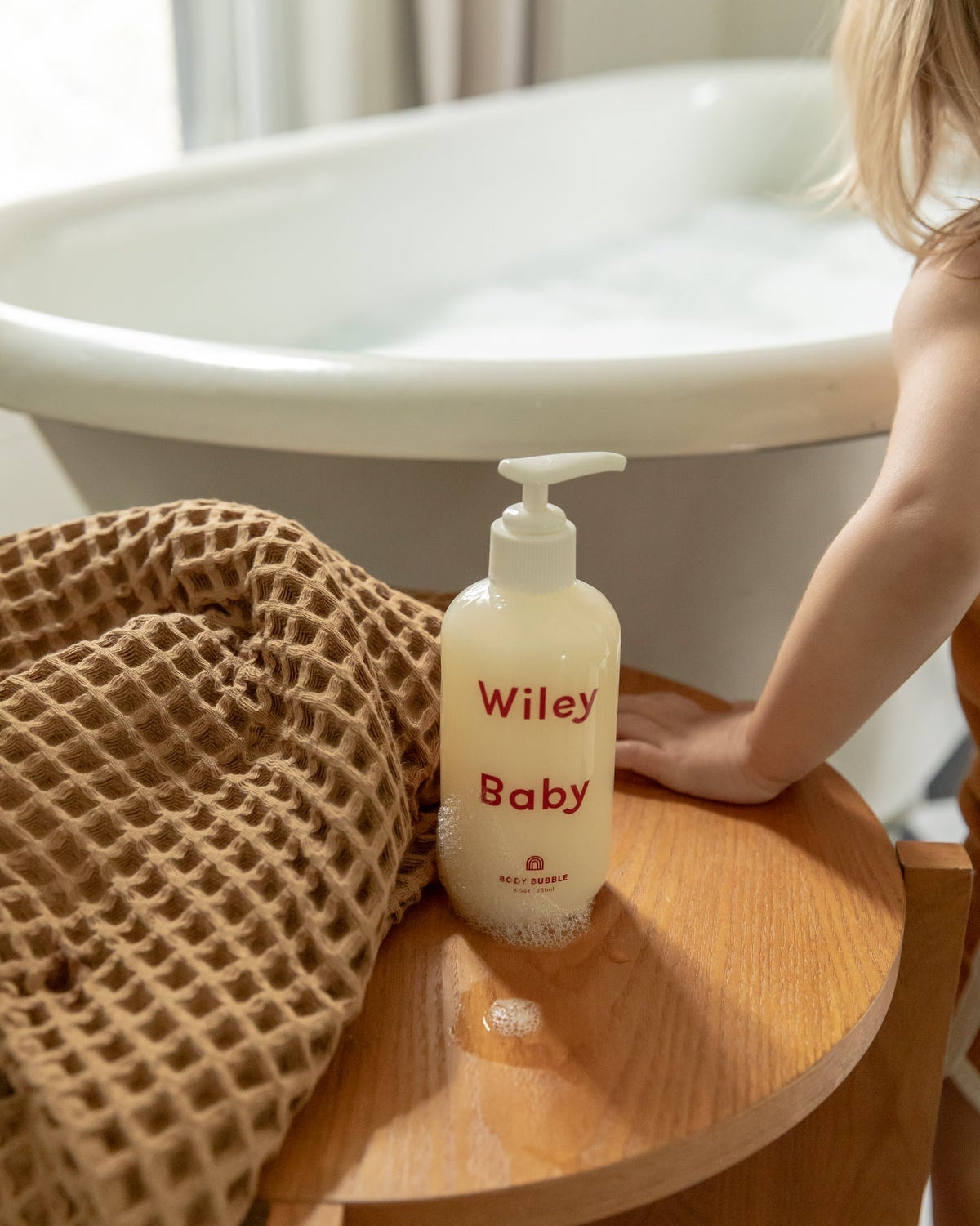 Wiley Baby bottle on a wooden stool next to a bathtub with a child's hand reaching out.