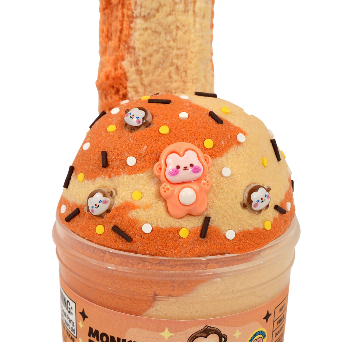 A novelty slime jar shaped like a donut, labeled 'MONKEY BREAD' on the front. The jar contains orange and beige marbled slime, topped with a beige donut-slice shape decorated with small monkey charms, white and black sprinkles, and a central pink monkey figure.