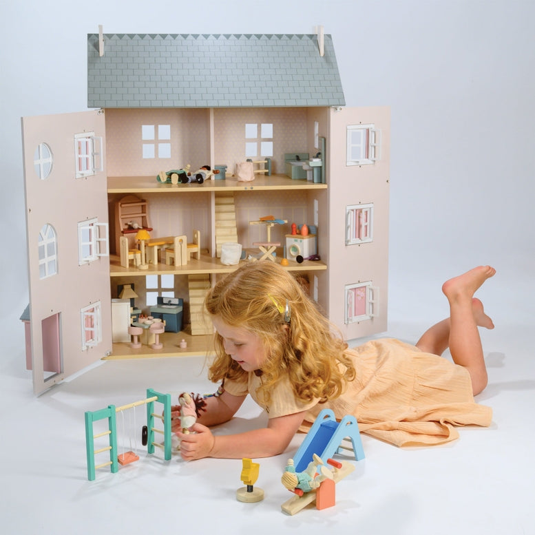 A young child with blonde hair wearing a beige dress lies on the floor playing with a large wooden dollhouse, alongside the wooden playground set featuring a see-saw, slide, climbing frame, swing, tire, and a bouncy bunny toy.