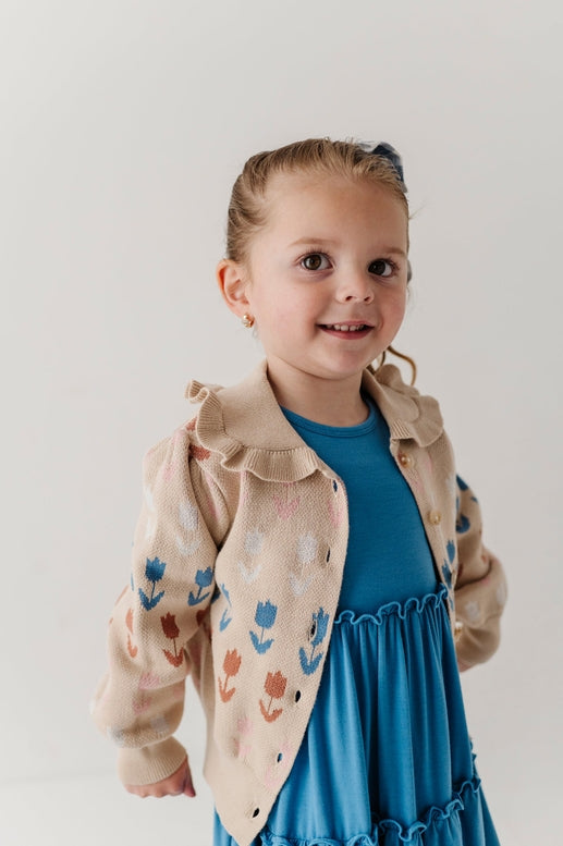 Young girl wearing a beige floral jacket over a blue dress on a plain background