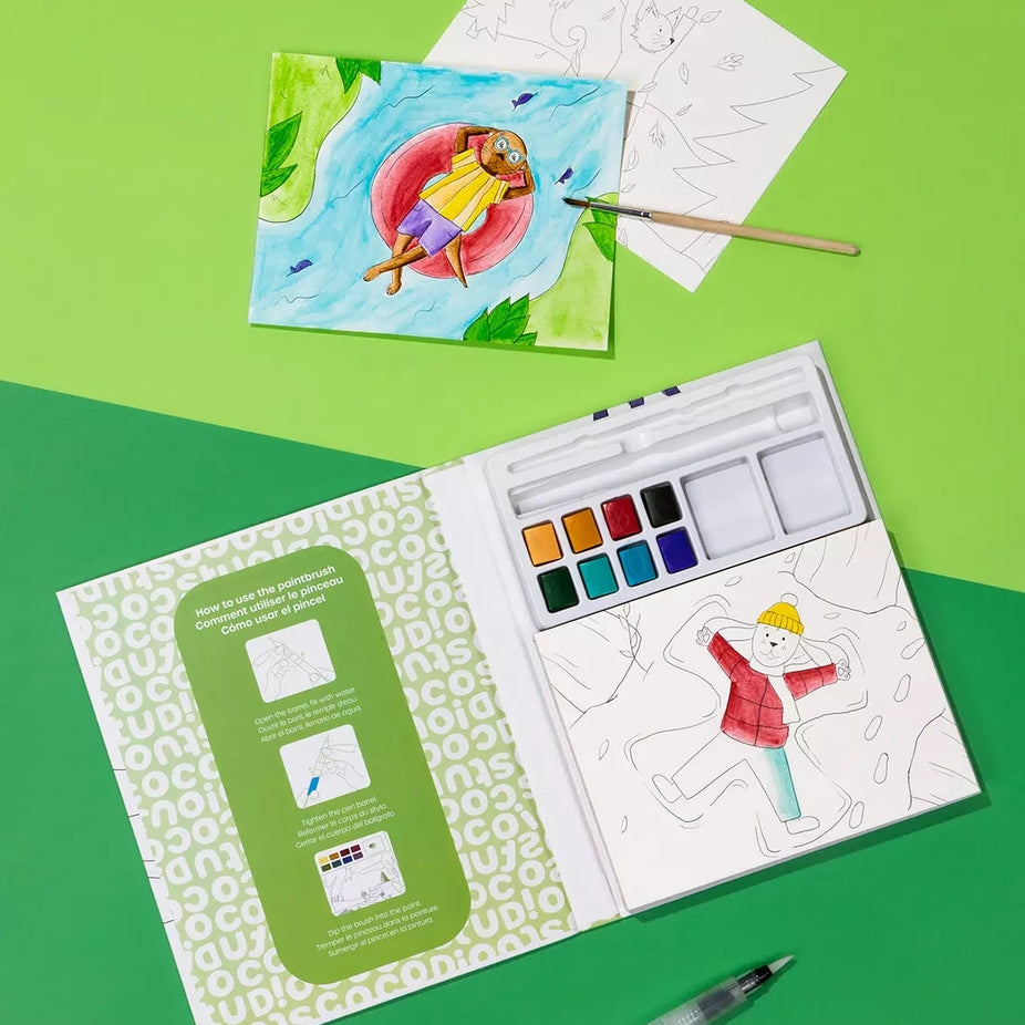 An open children's watercolor painting kit on a bright green background, with a painted sample card, a paintbrush, a pencil, and a water pen arranged around it. The kit includes a palette, paper, and printed instruction panel.